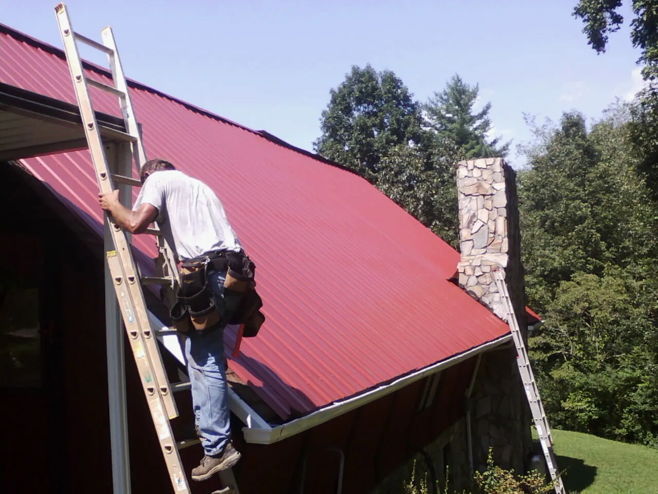 Roofer on a ladder beside a red metal roof and stone chimney — Soffit & Fascia Repair by Roofing Brothers in Placentia